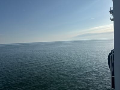 This media file shows a wide view of a calm, dark blue sea under a clear, pale blue sky with a few wispy clouds. The horizon line is sharp and separates the sea from the sky. The sun is not directly visible, but its presence is suggested by the bright, hazy band of light towards the horizon, indicating it is either early morning or late afternoon. On the right side of the image, a portion of a white railing and a dark object, possibly a life raft covered with a tarp, are visible. This suggests the photo was taken from a ship or ferry. The overall mood is serene and tranquil. There is no visible text in the image.