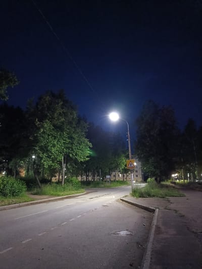 The image depicts a street scene at night in Chaikovsky, Russia, likely in a residential or semi-residential area. A road curves to the left, illuminated by a bright streetlamp. There are trees lining the road and a pedestrian crossing sign is visible near the streetlamp. Buildings with lit windows are present in the background, suggesting the presence of apartments or houses. The sky is dark blue, indicating it is nighttime. The overall impression is one of a quiet, ordinary street at night.