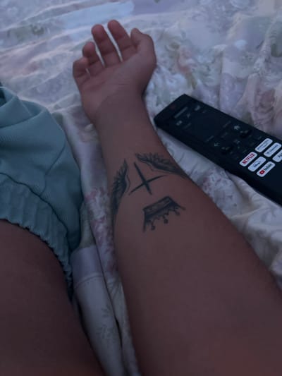 The image shows a person's arm with a tattoo of wings and a crown. A television remote control is visible on the bed next to the arm. The background appears to be a bed with patterned sheets. No specific location in Ognianovo, Bulgaria, is depicted.