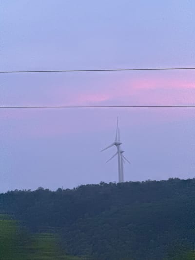 An outdoor, wide shot captures a tranquil, dusky scene in the city of Kuriany, Ukraine. A dark, undulating hillside dominates the lower portion of the frame, its surface obscured by dense foliage. The sky above is a soft gradient of lavender and pink, with a delicate band of pink clouds streaking across the upper right quadrant. Two white wind turbines stand in the distance, silhouetted against the twilight sky and perched on the crest of the hill. Their tall, slender structures and the sweeping blades of their rotors are clearly visible. Three thin, dark power lines stretch horizontally across the upper half of the image, dividing the sky and creating a subtle, linear pattern. The overall impression is one of peaceful, natural beauty.