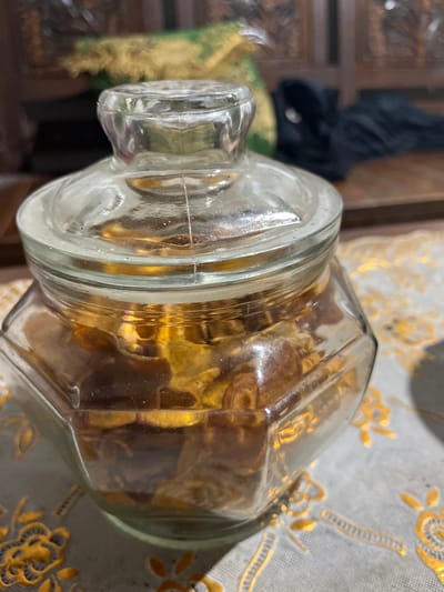 In Layansari, Indonesia, a close-up, eye-level shot captures a hexagonal glass jar filled with dried, amber-colored food items. The jar has a clear glass lid with a bulbous top. The jar rests on a white cloth with a golden floral pattern. The background is blurred, showing wooden furniture, a dark fabric, and a splash of green and yellow material, suggesting an indoor setting. The lighting is warm, creating reflections on the glass and highlighting the texture of the food inside.
