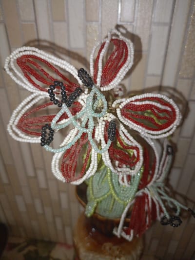 A beaded floral arrangement is displayed in a pot. The arrangement features white, red, and light green beads, with some black beads forming loops. The beads are meticulously crafted to resemble petals and leaves. The background consists of a tiled wall with a light beige color and a grid pattern. The lighting in the image is warm, casting soft shadows and highlighting the textures of the beads and the wall. The location of the object is in the city of Mytishchi, Russia.