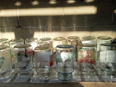 The media file shows a collection of empty glass jars arranged on a striped surface. The jars are of various sizes and shapes, some with remnants of labels still attached. The background is a rough, textured wall, possibly made of cinder blocks, with distinct shadows cast across it, indicating bright sunlight. This suggests it is daytime, likely outdoors, given the natural light and shadows. The jars appear to be either drying or stored. The overall scene is one of simple domestic storage or preparation. Some jars have blue-tinted rims or bases, and the labels are too blurry to read clearly, though some appear to have images of produce on them. There is no text that can be definitively read. The setting appears to be in a place with abundant sunlight, suggesting a warm climate. The image is focused on the objects themselves, with no people present. The location is identified as Теңги, Kyrgyzstan.