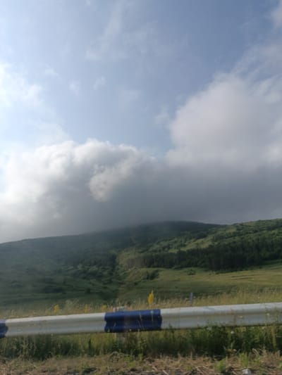 The image depicts a scenic view of a hilly landscape under a cloudy sky, with a roadside guardrail in the foreground. The guardrail is white with blue stripes. The hills are covered in green vegetation, appearing as a mix of grassy areas and scattered trees. The sky is filled with large, white and grey clouds, with patches of blue sky visible. The lighting suggests it is daytime, possibly late afternoon due to the soft light and some shadows on the hills. There are no people or animals visible in the frame. The setting appears to be a natural environment, likely a rural area, possibly along a road, given the presence of the guardrail.