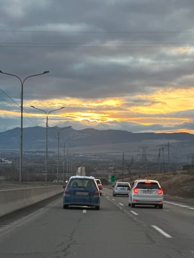 A cloudy sky with streaks of bright orange light flanks a roadway with cars. Three vehicles are visible, traveling on the right side of the road, a blue car in the foreground, followed by two white cars. Power lines and streetlight posts run along the edge of the highway. Hills and a cityscape are visible in the distance under a hazy sky.