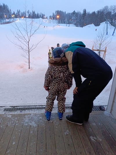 The image captures a winter scene at what appears to be a ski resort, viewed from a wooden deck or porch. In the foreground, two individuals are seen from behind. On the left, a small child is dressed in a full leopard-print ski suit with a fur-lined hood. They wear a dark blue ski helmet with partially visible white text that reads "PAUL FRANK" and bright blue ski boots. To the child's right, a taller individual, likely an adult, is leaning forward with their hands on their knees, appearing to be attentive to the child or something in front of them. This person wears a multi-colored ski jacket (dark blue, tan/cream, and teal/green) and dark snow pants, with black shoes or boots.
The immediate foreground features a wooden plank deck, partially covered in patches of snow and ice, and a metal grate or drainage cover running horizontally across the frame. Beyond the deck, the ground is covered in snow. A bare deciduous tree with thin branches stands prominently on the left side of the frame. In the mid-ground, skis and poles are propped up, and a wooden ski rack holding more skis is visible on the right.
The background reveals snow-covered ski slopes illuminated by several artificial lights, suggesting it is either dusk, dawn, or nighttime. Other skiers can be seen as small figures on the slopes. Coniferous trees (pines) covered in snow line the top of the hills and delineate the edges of the ski runs. Ski lifts are also visible further in the background. The sky is a pale, overcast blue-grey, contributing to the diffused lighting. The overall scene conveys a cold, snowy winter environment, likely a family outing or lesson at a ski area.