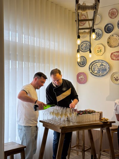 In Felgueiras, Portugal, two adult males are engaged in food and beverage preparation within an indoor commercial or event space. A male positioned on the left, wearing a white t-shirt and light blue jeans, is actively pouring a light-colored sparkling liquid from a green bottle into a row of multiple champagne flutes on a wooden high-top table. He has a dark bracelet on his left wrist. Immediately to his right, a male wearing a black chef's jacket with a tan diagonal stripe is cutting a large, rectangular cake with a silver knife. The cake is decorated with a top layer of mixed berries and has white, vertical, cylindrical elements along its sides. A collection of approximately 20 champagne flutes, many partially filled, are arranged on the table. Overhead, a decorative lighting fixture composed of a wooden ladder wrapped with rope suspends five bare Edison-style light bulbs. The background features a large window covered by vertical cream-colored blinds on the left and a white wall adorned with numerous decorative ceramic plates of various sizes and blue, pink, and white patterns on the right. A portion of a third individual's right shoulder and arm, wearing a white garment, is visible on the far right of the frame. A light-colored wooden stool is partially visible in the lower left foreground.