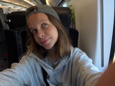 A young woman wearing a grey baseball cap and a grey hoodie is sitting on a train. She has freckles and a nose ring. The train appears to be a commuter train, with rows of dark seats visible in the background. There is a poster on the wall of the train car. The location is identified as Nemchinovka, Russia.