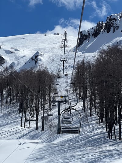 The image depicts a snowy mountain landscape with a ski lift in operation. The location is Katun Bačko brdo, Montenegro. The sky is blue with scattered clouds. The ground is covered in snow, showing ski tracks. There are bare trees alongside the ski slope, casting long shadows on the snow. The ski lift consists of chairs suspended from a cable. The ski lift structure and chairs are visible. The image is taken from a high vantage point, looking down the mountain.