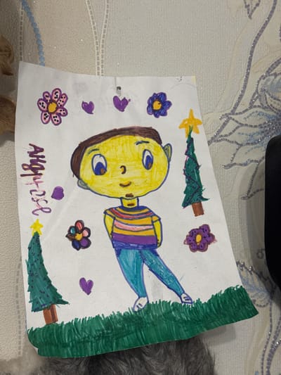 The image shows a child's drawing on white paper. The drawing includes a yellow-faced figure with brown hair, wearing a striped shirt, and blue pants. There are flowers and heart shapes drawn around the figure. At the top and bottom edges are stylized green fir trees. The drawing appears to be signed with the name "AHGPOSE". The background is a light-colored patterned surface with some indistinct objects and a section of dark gray fur or fabric. The image was likely taken in Chuprino, Russia.