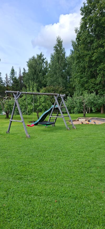 The image depicts a children's playground situated in a grassy park area, likely in Valma, Estonia. The playground features a wooden swing set with two swings, one red and one black disc-shaped swing. Attached to the swing set is a green slide. To the right of the swing set is a sandbox containing several toys, including what appears to be a blue bucket and spade. The background consists of lush green trees and a partly cloudy sky. The ground is covered in a well-maintained lawn. No people are visible in the scene.