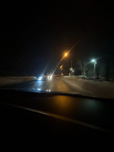 The image depicts a nighttime scene observed from the interior of a vehicle, looking forward through the windshield. The top portion of the dashboard is visible in the lower foreground. The environment is dark, suggesting night, with snow visible along the roadside.
A two-lane paved road stretches into the distance, illuminated by multiple streetlights and oncoming vehicle headlights. Several vehicles are approaching from the opposite direction, their headlights appearing as streaked bright white and blue light sources due to motion blur or camera exposure.
On the right side of the road, snowbanks line the edge. A prominent streetlight with an orange glow is visible, along with a secondary streetlight emitting a cooler, whiter light further down. Adjacent to the orange streetlight, a yellow diamond-shaped road sign, indicating a priority road, is mounted on a pole. Below this sign, a reddish-orange object or structure is present. Trees, partially obscured by darkness and snow, are visible behind the roadside elements. No individuals are explicitly visible. The scene is located in the city of Maksimkovo, Russia.