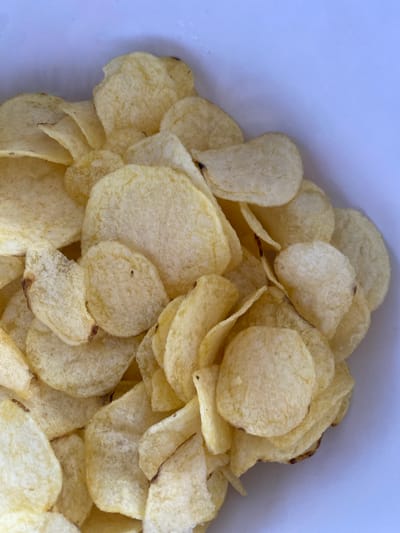 A close-up, overhead shot captures a pile of golden potato chips, their textured surfaces catching the light. The chips are irregularly shaped and some have browned edges, suggesting they are fried. The background is a solid, light blue-gray, which appears to be the inside of a container, possibly a bowl or bag. There is no discernible location in the city of Osek, Czechia, present in the image.