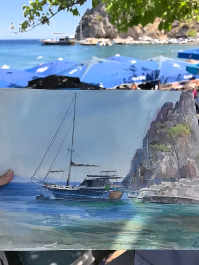 A watercolor painting depicts a serene harbor scene with two sailboats anchored near a rocky island. The foreground of the painting features the sailboats in calm, turquoise waters, with the larger sailboat prominently displaying its mast and rigging. The rocky island, rendered in muted tones with touches of green vegetation, rises majestically in the background. The sky above is a pale blue, suggesting a clear, sunny day. The painting is held by a hand in the lower left corner. The real-world background of the photograph shows a beach with numerous blue umbrellas, the sea with distant boats, and green foliage hanging from a branch in the upper left. This scene is likely located in Datça, Turkey, a region known for its beautiful coastline and natural harbors.