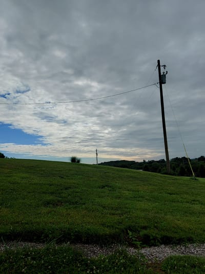 The image captures an outdoor scene, likely in Owingsville, United States, with a telephone pole dominating the left side of the frame. The pole is wooden and equipped with electrical components, including what appears to be a transformer, connected to overhead wires that stretch across the sky. The sky itself is filled with a mix of clouds, with some patches of blue sky visible, suggesting a partly cloudy day. The time of day appears to be either early morning or late afternoon, as indicated by the soft, diffused light.

To the right of the telephone pole, a lush green grassy field slopes downwards. A narrow strip of gravel or small stones runs along the edge of the field, bordered by more grass and some small white flowers. In the distance, a line of trees forms a dark green silhouette against the horizon, with the sky above them displaying a soft gradient of light, transitioning from a pale yellow or orange near the horizon to a lighter blue. The overall impression is one of a serene, rural landscape, possibly during a calm weather day. There is no visible text in the image.