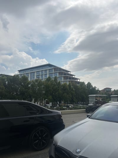 This media file depicts an exterior scene featuring a modern building with multiple stories and glass-paneled balconies, surrounded by lush green trees. In the foreground, the hoods and windshields of two cars are visible, one silver and one black. The silver car has a BMW logo on its hood. The scene is outdoors, with a cloudy sky overhead, suggesting it might be daytime but not necessarily bright sunlight. The buildings and dense foliage hint at an urban or suburban environment. There are no people visible, and the primary focus is on the architecture and the vehicles. The location context provided is Sabunchu Raion, Azerbaijan, which, combined with the visual elements, suggests a contemporary architectural development in that region. The overall impression is one of a modern, perhaps residential or commercial, area.