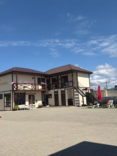 A two-story building with a brown tiled roof is visible under a bright blue sky with scattered clouds. The building has cream-colored walls with dark brown trim. A balcony runs across the second story, adorned with flower boxes containing pink and white flowers. The word "RECEPTION" is printed in black letters on the front of the building. To the right of the building, a metal staircase leads to the upper level. In front of the building, there is a paved courtyard with white outdoor furniture, including chairs and tables. To the far right, two lounge chairs with red cushions are set up on a patch of grass next to an inflatable pool. A red umbrella stands near the pool. The scene appears to be a hotel or guesthouse. The text "АМЗАРА. ПОЛИНА" is visible on the side of the building, indicating the location as Amzara, Abkhazia.