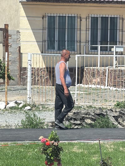 A middle-aged bald man with a prominent belly, wearing a blue and white striped tank top and dark pants, walks to the right along a paved path. He is in front of a yellow building with barred windows and a rusty metal fence. The building's facade is partially obscured by a shadow cast from the left. Behind the fence, the building wall is decorated with rough-hewn stones. To the left of the man, a small leafy plant grows near a rough, damaged wall. In the foreground, a patch of green grass is visible, with a small bush of red and pink flowers. The ground is a mixture of gravel and some dark debris. The overall setting appears to be a residential area in Balykchy, Kyrgyzstan.