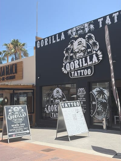 This image depicts the exterior of "Gorilla Tattoo," a tattoo parlor located in a sunny, commercial area, likely in La Oliva, Fuerteventura, Spain. The shop's facade is black with a distinctive white logo featuring a gorilla's head wearing sunglasses and a helmet, framed by palm leaves, with the numbers "20" and "24" incorporated into the design. The name "GORILLA TATTOO" is prominently displayed on the main sign and repeated on the shop's windows.

Two A-frame signs stand on the pavement in front of the shop. One sign provides key information: "GORILLA TATTOO FUERTEVENTURA walk-ins welcome OPEN EVERY DAY 10 to 10" and an Instagram handle "@gorillatattoofv." The other A-frame sign showcases a variety of small tattoo designs. A banner or awning above the main sign also bears part of the shop's name, "TAT."

The scene is brightly lit under a clear blue sky, suggesting a sunny day, with strong shadows visible on the light-colored pavement. Palm trees are visible to the left, indicating a warm climate. Below the tattoo shop, part of another business's sign reads "SA BEACH," implying a location near a beach. No people are visible in the photograph.