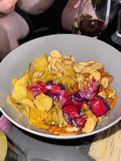 A close-up, eye-level shot shows a grey bowl filled with an assortment of snacks, including ruffled potato chips, crackers, pretzels, and small biscuit-like items. Several wrapped candies, two in red foil and two in purple foil, are nestled among the snacks. In the background, out of focus, are parts of people's legs, a can, and a wine glass containing red liquid. The scene appears to be indoors, possibly in a home or at an event. The image does not contain any information about the city of Jablonné nad Orlicí, Czechia.