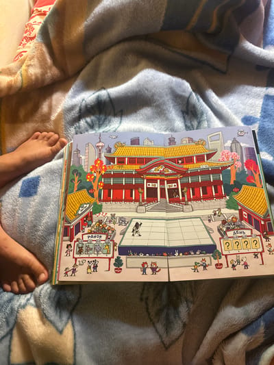 The image shows an open book with a colorful illustration of a traditional East Asian building with yellow tiled roofs, situated in a cityscape with modern skyscrapers. The book is lying on a soft, blue and tan patterned blanket. A child's bare feet are visible on the left side of the image, resting on the blanket near the book. The illustration depicts various cartoon animals dressed as humans, engaged in activities. In the foreground, there are signs with Russian text, including "РАНОК" (Morning) and "ДЕНЬ" (Day), with times and question marks, suggesting a schedule or event. There are also depictions of what appear to be sports activities on a court. The overall scene in the book is one of a bustling, possibly festive, event in a stylized urban environment. The location is not specified within the image itself.