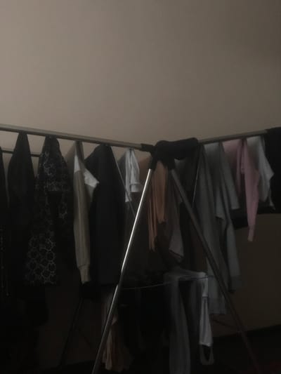 The scene depicts an indoor environment within Tashbulak, Uzbekistan, characterized by low ambient lighting. A collapsible metal clothing drying rack, constructed with silver-toned tubing and black plastic joints, stands centrally. Various garments are suspended from its horizontal bars, indicative of recent laundering and air-drying. Visible clothing items include dark shirts or jackets, one dark garment with a repeating light-colored pattern, a light beige long-sleeved top, several light grey and white shirts, and a prominent pink top. One light grey item exhibits vertical stripes. Additional light-colored clothing is discernible hanging on the lower tiers of the rack. The background is a plain, light-colored wall. No individuals are present.