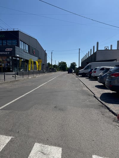 This is a medium-shot, eye-level, wide-angle photograph of a street scene in Sarny, Ukraine. The image is oriented vertically, as if captured while holding a phone in portrait mode. The right side of the frame shows a paved road with a white line marking the center. The left side of the frame features a modern building with a dark gray facade and large glass windows. Several yellow banners with black text hang in front of the building. Partially visible text on one banner reads "ECONOM CLASS". Above the building, construction is underway, with concrete pillars and scaffolding visible.
In the foreground, a shadow cast by an unseen object falls across the road. Further down the road, parked cars are visible along the sidewalk. The sky is a clear, bright blue, suggesting a sunny day. The overall impression is one of a commercial area on a bright, possibly warm day. The presence of construction indicates ongoing development in the area. The scene appears quiet, with no people prominently featured.