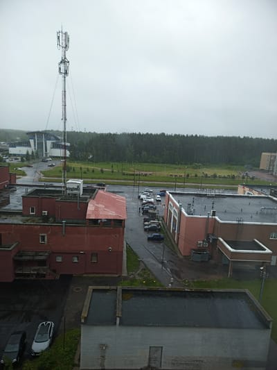 An aerial view captures a cloudy day in Lomonosov, Russia, focusing on a mix of industrial buildings, a cell tower, a sports complex, and a surrounding forest. In the foreground, a two-story red brick building with a flat roof and a separate section with a corrugated red roof occupies the lower left and center of the frame. To the right of this building is a larger, modern pink and white commercial building. A parking lot stretches between these structures and a road, with several cars parked, including a black sedan and a silver hatchback in the lower left foreground.

Further back, across a wet asphalt road, lies a spacious sports complex with a distinctive blue and white architectural design. Adjacent to the sports complex is a dense forest, forming a dark green backdrop that extends to the horizon. Several streetlights are visible in the green spaces and along the road.

Dominating the mid-ground is a tall, multi-tiered cell tower with antennas, situated on a base structure of a building. The overall scene depicts an urban fringe environment characterized by utilitarian architecture, recreational facilities, and natural woodland, under an overcast sky.