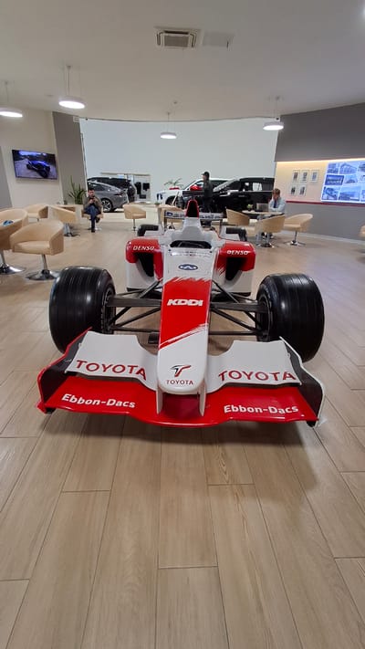 This is a photograph of a Toyota Formula 1 race car model displayed indoors, likely in a showroom or exhibition space. The car is predominantly white with red accents and features sponsor logos such as "TOYOTA," "DENSO," "KDDI," and "Ebbon-Dacs." The setting has light-colored wooden plank flooring.

In the background, several people are visible. Some are standing near other vehicles, possibly cars, while others are seated in chairs. One person is leaning over a black car. There's a television screen mounted on the wall displaying an image. The overall lighting suggests it is daytime, and there are no obvious cues about the weather. The atmosphere appears casual, with people browsing or waiting. The location is identified as Novogorelovo, Russia.