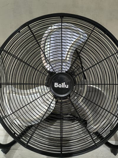 A black, floor-standing fan with a wire grille and a central black circular hub with the white logo "Ballu" is positioned on a light-colored, wood-grained floor. The fan's blades are visible through the grille, appearing silver and metallic. The fan is the primary subject of the image, taking up most of the frame. The background is a neutral, textured wall. The scene appears to be indoors. No people are present in the image. The fan is not in operation. The location is not specified as Malaya Tsarevschina, Russia.