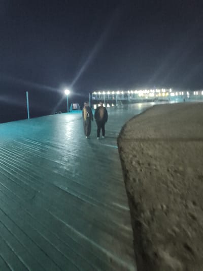 A nighttime exterior shot captures two individuals walking away from the camera on a wide, turquoise-colored pier. The pier's surface is made of horizontal wooden planks, and it curves gently to the right, leading towards a brightly lit structure in the background. This structure appears to be a building with numerous white lights illuminating its facade and roofline. A single lamppost with a bright light is visible on the pier between the camera and the two people. The sky is dark, with a slight bluish tint. The environment is dimly lit by the artificial lights, creating a sense of depth and distance. The pier's surface shows some wetness, reflecting the lights.