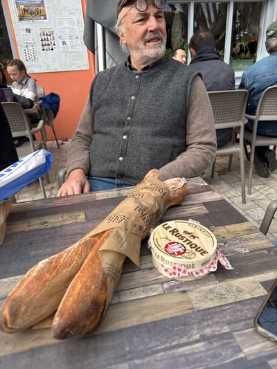 The image depicts an outdoor dining scene at a cafe in Cucuron, France. A man, approximately 60-70 years old with a grey beard, is seated at a rectangular table made of a material resembling dark wood planks. He wears a dark grey wool vest over a lighter brown long-sleeved shirt and blue jeans, with round spectacles perched on his head. He is looking off to the right, slightly upward, with a faint smile. His left hand rests on the table.
On the table in front of him are two baguettes, partially wrapped in brown paper bearing the text "L'Estival" and other French words. To the right of the baguettes is a round, wooden box of "LE RUSTIQUE CAMEMBERT" cheese, wrapped in a red and white checkered cloth. A small blue and white paper bag is visible to the far left on the table. A smartphone, partially visible, rests on the table's bottom right corner.
In the background, other patrons are seated at similar outdoor tables. To the left, a young girl with dark hair, wearing a striped top, is visible. Behind her, a white bulletin board with several printed posters, some featuring small photographs, is affixed to an orange wall. To the right of the man, another patron is visible from behind, wearing a dark jacket, and further right, another person in a denim jacket. A building with large glass windows and hanging plants is visible behind these patrons. The ground beneath the tables appears to be paved.