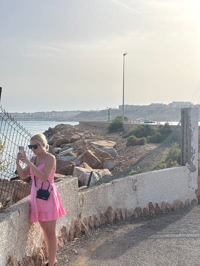 A blonde woman wearing a pink polka dot dress and sunglasses is standing by a stone wall, holding a smartphone and appearing to take a photograph or record a video. She is near a wire fence and a rocky shoreline. In the background, the sea is visible, with a distant cityscape and several boats on the water. The scene takes place outdoors on a clear day with bright sunlight. The environment suggests a coastal location. The city of Urbanización Cabo Roig, Spain, is mentioned as the location.