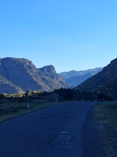 A narrow, dark asphalt road curves through a mountainous landscape under a clear blue sky. The road is bordered by a steep, rocky mountain slope on the left, covered in patches of green vegetation and some darker, larger rocks. The mountain peaks are rugged and layered, showing distinct geological formations. On the right side of the road, there is a grassy embankment that leads up to more rocky terrain and sparse vegetation. The sun appears to be low in the sky, casting long shadows and creating a sense of early morning or late afternoon. There are a few white poles or markers visible along the side of the road. The overall scene suggests a remote and natural setting, possibly a scenic route for driving or cycling. No people are visible in the image. The location is identified as Shatin, Armenia.