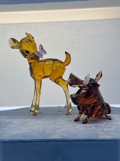 A close-up shot depicts two glass figurines against a backdrop of white and blue fabrics. The foreground figure is a golden deer posed mid-leap, adorned with small purple butterflies on its head. Behind it, a brown, glass bull figure is visible. The location is likely indoors, and though specifics regarding Spreitenbach, Switzerland cannot be determined from the visual elements, the scene likely represents a decorative display.