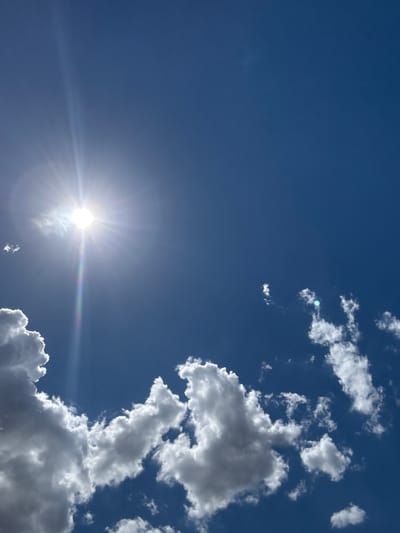This is a vertical, high-angle shot of a clear blue sky with scattered white clouds. The sun is prominently visible in the upper left quadrant of the image, emitting bright rays and a rainbow-like lens flare. Some of the clouds are illuminated by the sun, appearing bright white, while others are in shadow, showing darker gray tones. There are no people or man-made objects visible in the image. The scene depicts a sunny day in an outdoor environment. Without additional information, the specific location of Бурсоль, Russia cannot be confirmed from the visual content alone.