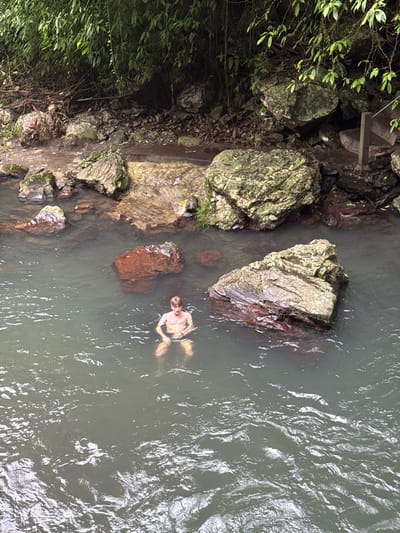 An overhead shot shows a man floating in a river or stream, partially submerged. Large, mossy rocks line one bank, with dense green foliage growing nearby. Smaller, reddish-brown rocks are visible in the water near the man. The water is murky, with visible ripples and currents on the surface. The man is pale and appears to be relaxing in the water.