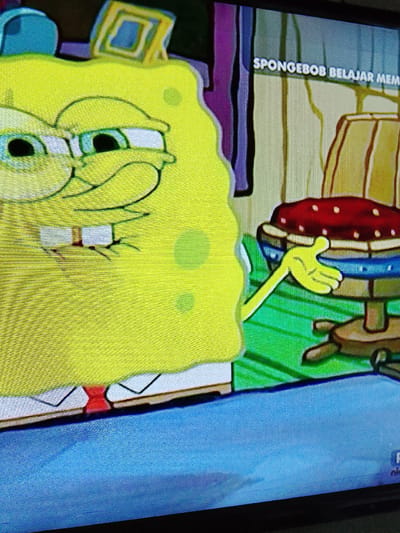 The image shows a close-up of a television screen displaying a scene from the animated show SpongeBob SquarePants. SpongeBob, a yellow sea sponge, is partially visible on the left side of the frame, with his signature square pants and red tie. He appears to be looking off to the side with a concerned expression, his eyes wide and his mouth slightly open. In the background, a wooden chair with a red, polka-dotted cushion is visible. To the right of SpongeBob, a green surface and vertical striped pattern suggest an indoor environment. At the top right of the screen, text in white letters reads "SPONGEBOB BELAJAR MEM". The image is taken from an angle, and the screen exhibits a grid-like pattern, indicating it is a photograph of a display. There is no information in the image that places it in Merbopuntong, Indonesia.