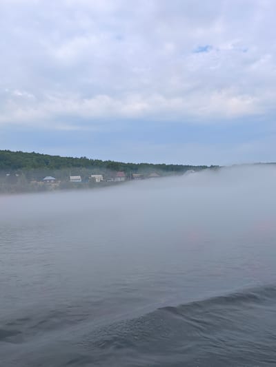 A wide, calm body of water, likely a river or lake, fills the foreground of the image. The water's surface is dark and slightly rippled, with a subtle wave forming on the right side. In the middle ground, a dense, low-lying fog bank partially obscures a shoreline. Beyond the fog, a treeline of dark green foliage stretches across the horizon, punctuated by a small village. Several houses with pitched roofs and varying colors are visible nestled amongst the trees. The sky above is overcast with a pale blue hue and soft, scattered clouds. The scene appears to be located in the village of Атаманово, Russia, given the context of the provided hints. No people or specific actions are depicted.