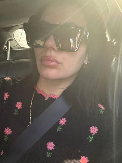 A female individual is depicted from the chest up, seated inside a vehicle. She wears oversized, dark rectangular sunglasses that obscure her eyes. Her lips are coated with a pink gloss, and her dark hair is visible, parted on the left. She is dressed in a black knitted garment adorned with embroidered pink daisies and green stems, featuring a pink scalloped trim at the neckline. A thin, gold-colored chain necklace is visible around her neck, and a small gold-colored hoop earring is present in her right ear. A dark-colored seatbelt crosses her torso diagonally. The background of the vehicle interior is indistinct due to significant sunlight entering from the upper right, creating a bright flare. Reflections visible on the sunglasses suggest an outdoor environment. This scene is located in Chew Magna, United Kingdom.