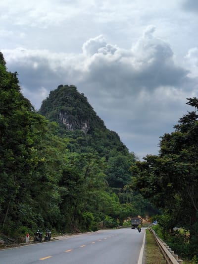 An overcast sky looms over a lush, green mountainous landscape in Sơn Dương Commune, Vietnam. A paved road with yellow dashed lines runs diagonally from the lower left to the middle right of the frame, bordered by dense foliage on both sides. In the foreground, on the left side of the road, two motorcycles are parked. Further down the road, a dark truck is moving away from the viewer, followed by a motorcyclist. The mountain in the background is covered in thick vegetation, with a rocky outcrop visible on its peak. Signs are present along the right side of the road.