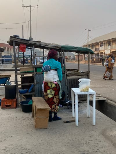 A woman wearing a turquoise top, white wrapper, and patterned skirt stands at a roadside stall. She has a baby strapped to her back. A white plastic bucket filled with yellow items rests on a white plastic table beside her. The stall is cluttered with various containers and supplies. The scene takes place on a street with buildings visible in the background. Another woman is further up the road, out of focus.