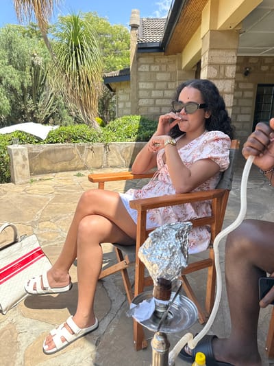 The scene depicts two individuals engaged in leisure activities on an outdoor stone patio in Olkaria ward, Kenya, under a clear daytime sky. A woman, with dark curly hair and wearing dark sunglasses, a light-colored floral short-sleeved top, white shorts, and white flat sandals, is seated in a wooden-framed canvas chair. Her legs are crossed at the knee. She holds a small, slender object to her mouth with her right hand; a gold-colored watch is visible on her left wrist. To her right, a second person, identified by dark skin tone and wearing black slide sandals and dark shorts, is partially visible. This individual holds a white flexible hose from a hookah with their right hand, while their left hand holds a smartphone. A silver-colored hookah, with its bowl covered in aluminum foil, is positioned on the stone patio floor between the two individuals. To the left of the woman's feet, a white tote bag with red stripes and "GUESS" branding rests on the patio. In the background, a light-colored stone building with a dark tiled roof and wooden eaves is visible, accompanied by lush green vegetation including palm-like trees, cacti, and shrubs. A clear glass containing liquid is placed on a low stone wall to the left of the woman.