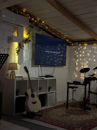 The image depicts an interior room, possibly an attic space, with a pitched roof lined with wooden beams. The walls are white brick. A television screen displays the message "check TX's input signal." Below the television is a white decorative cross adorned with floral elements, and a small table lamp is nearby. To the right of the television, a white shelving unit contains plants and other items. A white acoustic guitar rests on a stand adjacent to the shelving unit. A drum kit is positioned in the upper right corner of the image. A patterned rug partially covers the floor in the background. The ceiling and walls are decorated with string lights. An air conditioning unit is attached to the wall below the roof. The scene likely takes place in a residential or studio setting. The location is specified as Zgrade, Montenegro.