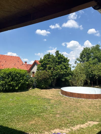 This is a landscape photograph taken outdoors on a sunny day. The scene is a backyard with a grassy lawn. In the foreground, the edge of a wooden structure, possibly a roof or overhang, is visible on the left. Beyond this, a red-tiled roof of a house and lush green bushes are present. Further back, a larger building is partially visible, surrounded by trees. On the right side of the image, a large, circular object with a wood-grain pattern around its circumference and a white interior is partially in view, suggesting it might be a pool or a similar structure. The sky is blue with scattered white clouds. The overall impression is that of a peaceful, residential outdoor space during daytime. No people or animals are visible, and no text is present in the image. The time of day appears to be afternoon, given the bright sunlight and shadows. The weather is clear and sunny.