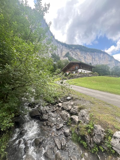 A wooden house with a red umbrella on a patio sits on a grassy hillside in Lauterbrunnen, Switzerland. A paved path winds up the hill toward the house. In the foreground, a stream flows over rocks, bordered by lush green foliage. The imposing Swiss Alps rise dramatically in the background, with a sheer cliff face and a lush green forest atop it. The sky is partially cloudy.