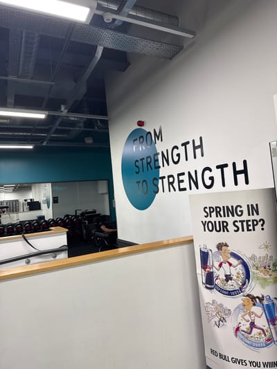 The image shows the interior of a gym, likely PureGym, as indicated by the logo reflected in the mirror. The gym features a weights area with rows of dumbbells and exercise machines, including a person on a machine in the background. A motivational phrase, "FROM STRENGTH TO STRENGTH," is prominently displayed on the wall in large black lettering within a teal circle. To the right of this, a Red Bull promotional banner is visible with illustrations of people running and text that says "SPRING IN YOUR STEP?" and "RED BULL GIVES YOU WIiiN[G]S". The overall environment appears clean and well-lit, with industrial ceiling fixtures and ductwork overhead.