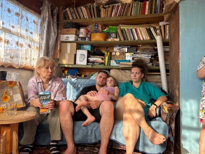 In the city of Маркидоновка, Russia, a family of four is seated on a blue sofa in a rustic, cluttered room. The room is lined with wooden shelves filled with books and various items. The family consists of an elderly woman on the left, a man holding a young girl on his lap in the center, and a young woman on the right. The elderly woman, with blonde hair, is wearing a patterned shirt and grey pants, and is holding a carton of juice. The man, in a black t-shirt and shorts, has his arm around the girl, who is wearing a pink dress. The young woman, in a teal top, is seated with her legs crossed. Sunlight streams in from a window on the left, illuminating the scene. A small wooden table with a glass and a pitcher is positioned to the left of the sofa. The overall impression is one of a humble and lived-in domestic setting.