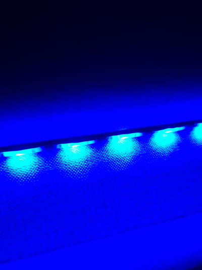 The image shows a close-up, angled view of a series of bright blue LED lights illuminating a textured surface, likely fabric or mesh. The lights are arranged in a horizontal line, casting distinct circular patterns of light and shadow on the surface below. The background is a deep, dark blue, creating a high-contrast effect. There are no people or animals visible in the image. The scene is abstract and does not provide any specific clues about its location in Evere, Belgium, or any other urban environment.