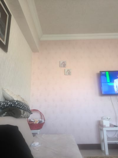 This image presents an interior view of a room, likely a living room or bedroom, located in Etimesgut, Turkey. The perspective suggests the photo was taken from a seated or lying position on a sofa or bed.

The room features walls covered in wallpaper: the left wall displays a subtle light-colored pattern, while the main wall to the right has a light pink background with a delicate floral or vine motif. The ceiling, visible at the top, is white and features decorative molding.

On the right side of the main wall, a flat-screen television is mounted. It is turned on, displaying a broadcast with the "TRT 1" logo visible in the upper left corner of the screen. A person wearing a white suit is partially visible on the television. Below the TV, a small white side table holds an electrical outlet, a white box, and several cables.

To the left of the scene, part of a sofa or bed is visible, covered with light-colored bedding. Stacked on this furniture are multiple pillows, including one with a black and white patterned cover, and a dark blanket. A white power adapter and cable rest on the bedding in the foreground. Further to the left, a red woven basket sits on a small white table. The basket contains various small boxes, with "Ecopirin" visible on one of them. Two small square framed pictures, depicting cherubs or similar angelic figures, adorn the light pink wall above the basket.

The overall scene suggests a calm, domestic setting, possibly captured during daytime, with no direct activity beyond a television broadcast.